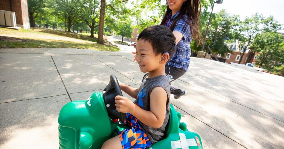 A young child, smiling, rides on a green toy with an adult, also smiling, running alongside.