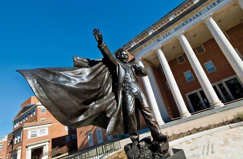 A bronze statue of Frederick Douglass, one arm extended with palm pointed toward the sky and his mouth open in speech, in front of the columns of the Hornbake Library entrance