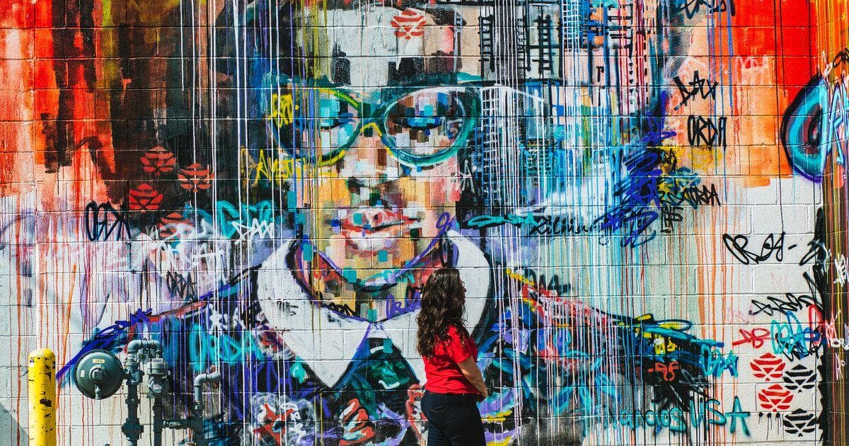 A mural painted in vibrant colors depicts a person's face with multiple shades of color and graffiti style lettering surrounding on all sides
